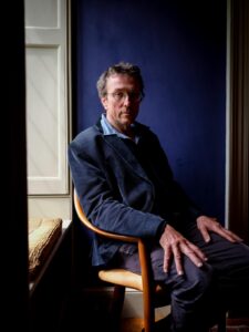 Man seated indoors with a blue wall background, dressed casually in dark jacket and glasses, capturing tranquil coastal ambiance.